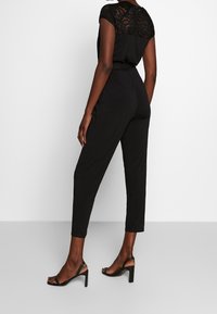 Une combinaison noire avec un dos en dentelle, une silhouette ajustée, des jambes fuselées et des manches courtes, accompagnée de sandales à talons noires.