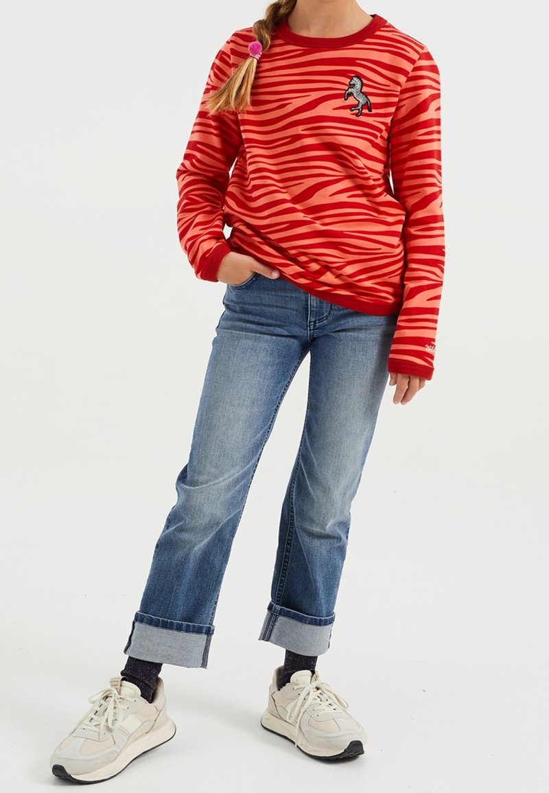 Enfant portant un t-shirt à manches longues rayé rouge et orange avec un patch cheval, un jean bleu retroussé, des chaussettes foncées et des baskets blanches.