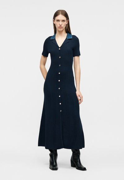 Γυναίκα που φοράει μπλε ναυτικό ριμπ φόρεμα με κουμπιά, κοντά μανίκια, γιακά από τζιν και μαύρα μποτάκια με τακούνι, στέκεται μπροστά σε απλό φόντο.