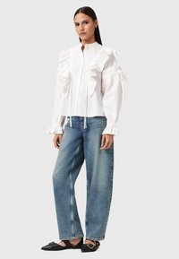 Kvinna står iklädd en vit volangblus med långa ärmar, lösa blå jeans och svarta spetsiga ballerina skor med spännen, framför en enkel bakgrund.