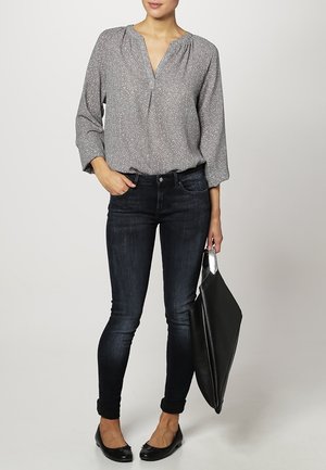 Blouse gris clair à motif avec un col en V et un tissu doux, associée à un jean skinny foncé et des ballerines noires, tenant un sac fourre-tout noir.