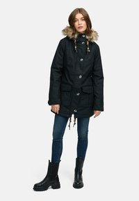 Svart parkas med huva i fuskpäls, stora framfickor, knappstängning, justerbara dragsnören och textilstruktur. Bärs med mörka jeans och stövlar.