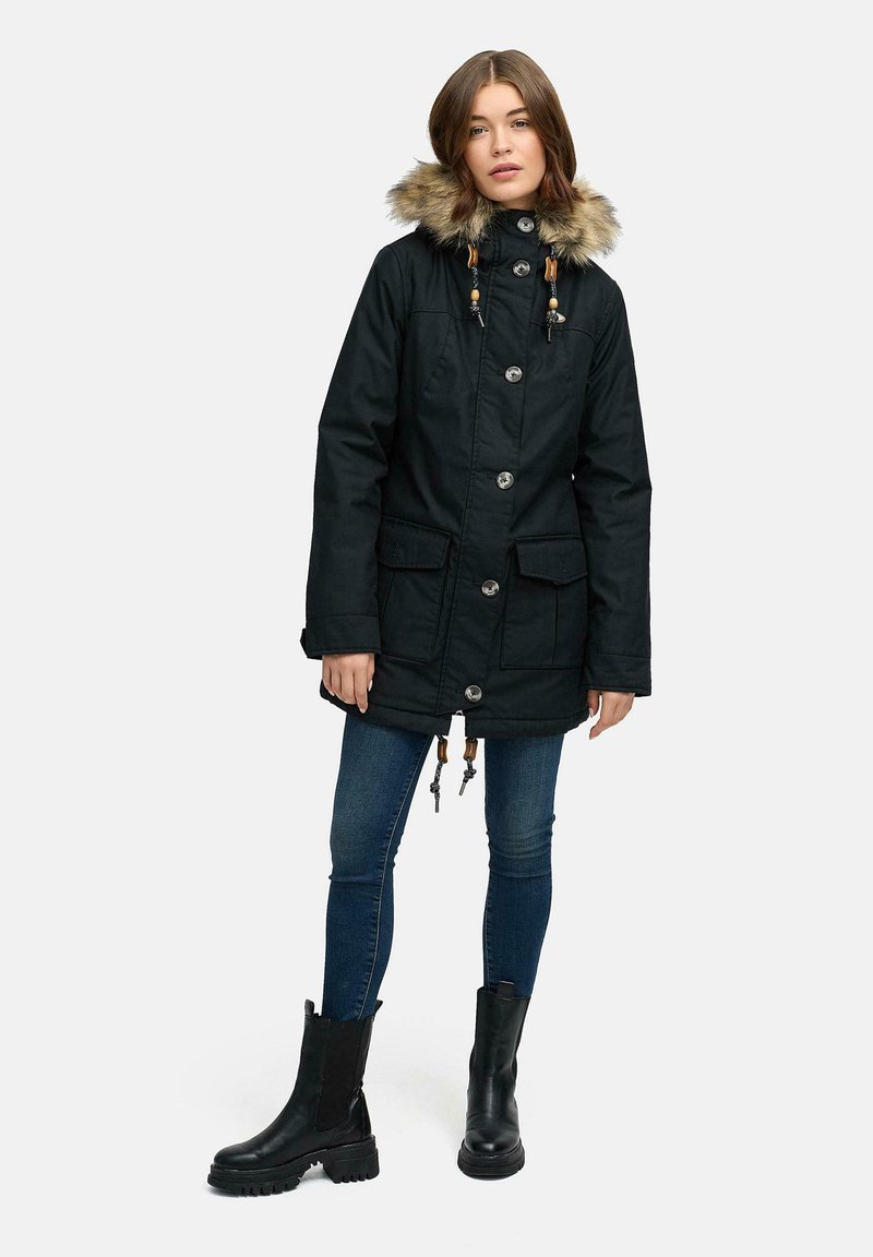 Svart parkas med huva i fuskpäls, stora framfickor, knappstängning, justerbara dragsnören och textilstruktur. Bärs med mörka jeans och stövlar.