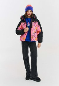Nafúkaná ružová bunda s kvetinovými a okom podobnými vzormi, čierne rukávy, zips na prednej strane, spárovaná s modrou košeľou, rozšírenými džínsami a čiernymi topánkami.