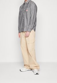 Man met een grijs overhemd met knopen en borstzakken, beige cargobroek en witte sneakers, staand tegen een effen witte achtergrond.