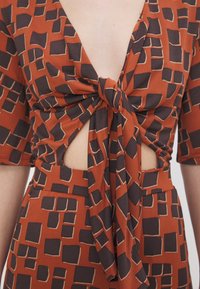 Haut à motifs géométriques orange rouille et noir avec manches mi-longues, présentant un nœud sur le devant et un détail découpé au niveau du ventre.