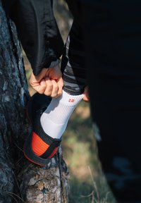 Czarno-pomarańczowe buty sportowe z teksturowanym wzorem, w zestawieniu z białymi skarpetami sportowymi z małym czerwonym wzorem, w tle drzewa.