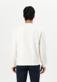 White crewneck sweatshirt with a soft texture, ribbed cuffs and hem, and a relaxed fit, worn with dark navy pants, viewed from the back.