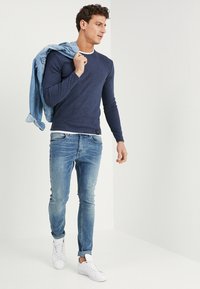 Granatowa koszula z długim rękawem, jasnoniebieskie dżinsy i białe trampki. Dżinsowa kurtka zarzucona na ramię. Prosty design i luźny krój.