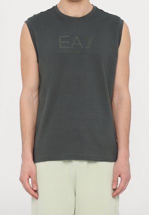 Hombre con una camiseta sin mangas Emporio Armani EA7 de color gris oscuro y pantalones de color claro, mostrado desde los hombros hasta las caderas.