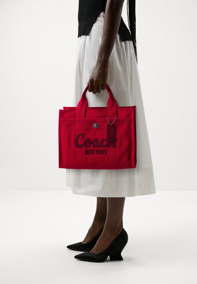 Personne portant une jupe blanche et un haut noir, tenant un sac cabas rouge Coach New York et portant des talons noirs pointus.