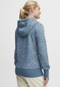 Blå hoodie i mjukt material med ett spritt cirkelfloralmönster i rosa och vitt. Har en dragsko i huvan och ribbade ärmslut.