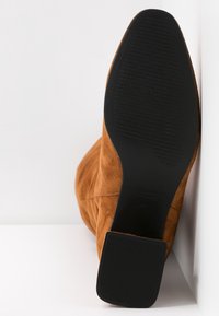 Botín de ante marrón con punta cuadrada y suela de goma negra texturizada. Presenta un diseño minimalista con líneas limpias y sin herrajes visibles.