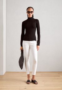 Black long-sleeve top, white fitted jeans, black slide sandals, holding a black bag. Smooth fabric, minimalistic design, neutral colors.