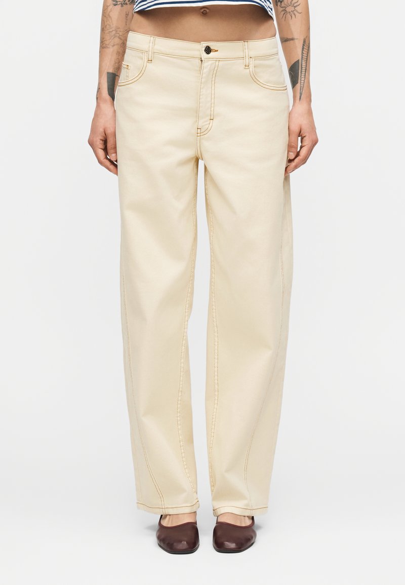 Άτομο που φοράει ψηλόμεσο, άνετο μπεζ παντελόνι με κουμπί και φερμουάρ, συνδυασμένο με σκούρα καφέ παπούτσια τύπου slip-on και ριγέ κοντό τοπ.