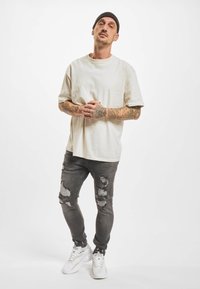 Lysebeige overdimensjonert t-skjorte, grå slitte skinny jeans og hvite joggesko. Tatoveringer synlige på armene. Minimalistisk design, avslappet passform.