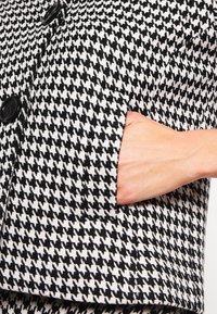 Kabát s černobílým vzorem houndstooth, se dvěma knoflíky a boční kapsou. Texturovaná tkanina a strukturovaný design.