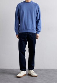 Ljusblå sweatshirt med rund hals och logga på bröstet, kombinerad med marinblå byxor och vita sneakers med bruna sulor. Mjukt tyg, ledig passform.