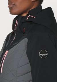 Gros plan d'une personne portant une veste matelassée noire et grise avec une fermeture éclair rose et des cordons, ainsi qu'un patch rond avec le logo Icepeak sur la manche.