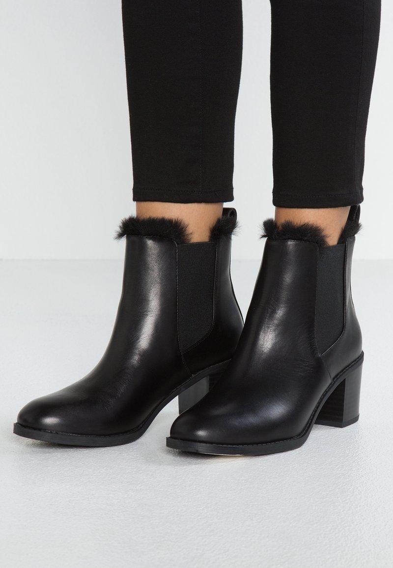 Botas de cuero negras hasta el tobillo con tacones de bloque y ribete de piel de cabello, llevadas con pantalones negros ajustados sobre un fondo de suelo claro.