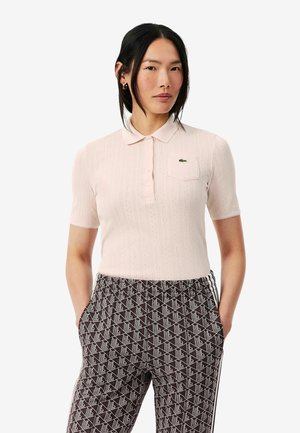 Mujer con cabello negro que lleva una camisa polo de manga corta de color rosa pálido y pantalones estampados en negro y blanco con las manos en los bolsillos.