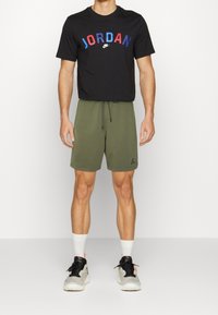 T-shirt noir avec "JORDAN" en rouge et bleu, associé à un short vert olive. Baskets noires et grises avec des accents rouges et chaussettes blanches.