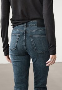 Person wearing dark gray long-sleeve top and high-waisted blue jeans, standing with hands relaxed by sides against plain background.