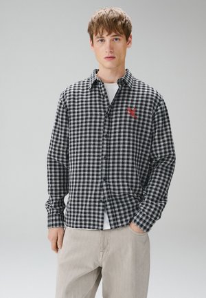 Ung mand iført en sort- og hvidternet skjorte med et rødt broderet design, beige bukser og en hvid undertrøje, stående mod en ensfarvet baggrund.