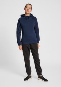 Granatowa bluza z kapturem, ze sznurkiem i długimi rękawami, zestawiona z czarnymi spodniami dresowymi i czarnymi butami sportowymi.