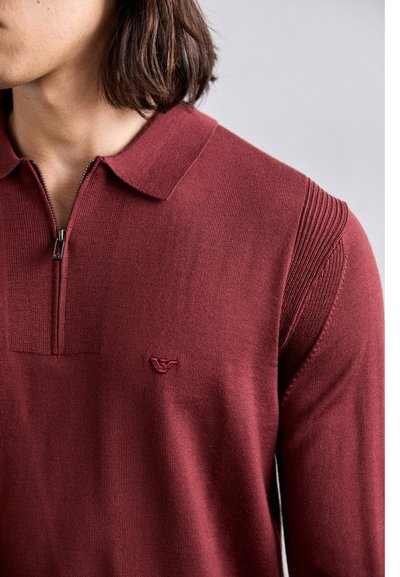Polo di color bordeaux con colletto a zip, modelli a trama sulle spalle e un piccolo logo ricamato sul petto. Tessuto a maglia, design aderente.