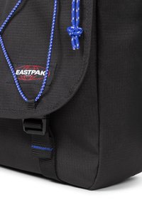 Musta reppu, jossa on teksturoitu pinta, sinivalkoinen naru, nappisulku ja etupuolella Eastpak-logo.