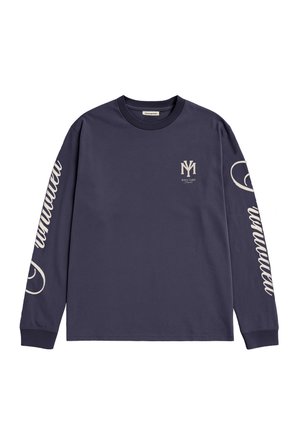 Homme portant une chemise bleu marine à manches longues avec des motifs blancs, une écharpe rouge à motifs et une casquette bleu marine avec le texte « Champagne Courtside Prohibited ».