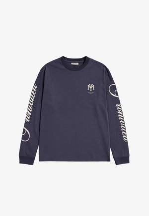 Homme portant une chemise bleu marine à manches longues avec des motifs blancs, une écharpe rouge à motifs et une casquette bleu marine avec le texte « Champagne Courtside Prohibited ».