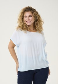 Camisetta corta a maniche corte di un blu chiaro semi-trasparente con collo rotondo e fondo elastico. Presenta una texture morbida e una vestibilità rilassata. Indossata con jeans scuri.