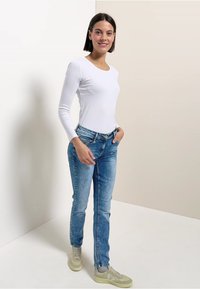 Witte lange mouwen overhemd, blauwe spijkerbroek en lichte sneakers. De spijkerbroek heeft een klassieke pasvorm en subtiele vervaging; het shirt heeft een ronde hals.