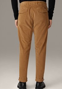 Persoon in camel-kleurige ribfluwelen broek met opgerolde broekspijpen en zwarte schoenen, gezien van achteren tegen een effen achtergrond.