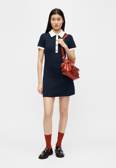 Woman wearing a navy dress with white collar and cuffs, holding a red handbag, paired with red socks and black loafers, standing against white background.