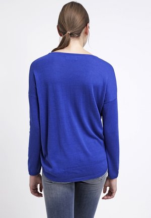 Frau mit braunen Haaren zu einem niedrigen Pferdeschwanz gebunden, trägt einen langärmeligen royalblauen Pullover und graue Jeans, steht mit dem Rücken vor einem weißen Hintergrund.
