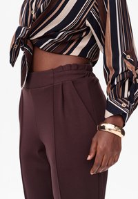 Vrouw met een hoge taille bruine broek en een gestrikte, gestreepte lange mouwen blouse, geaccentueerd met een grote gouden armband om de pols.