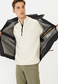 Witte gebreide half-zip trui, gedragen onder een donkere jas met grijze voering. Kenmerkt zich door een zak met rits en een logo, gecombineerd met lichtgroene broek.