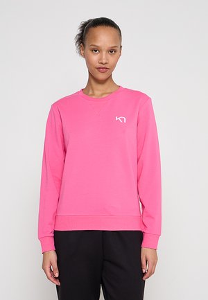 Donna che indossa una felpa a maniche lunghe di un rosa acceso con un piccolo logo bianco "KN" sul petto, in piedi davanti a uno sfondo chiaro e uniforme.