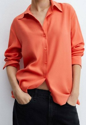 Personne portant une chemise corail amples à manches longues retroussées et un pantalon noir, les mains dans les poches.