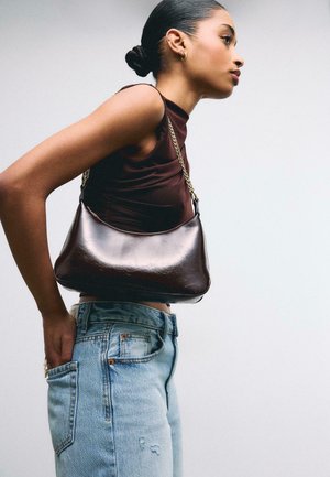 Ženska v brezrokavniku rjave barve in svetlo modrih kavbojkah nosi torbico s trakom iz zlatega verižnega materiala, gleda v stran na enobarvni ozadju.