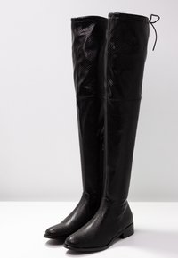 Botas negras por encima de la rodilla hechas de material sintético texturizado. Presentan un cierre lateral, tacón plano y puntera redondeada, con un caña ajustada.
