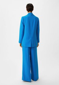 Heldere blauwe op maat gemaakte kostuum met een lange blazer en brede broekspijpen, gemaakt van een gladde stof met een strakke, minimalistische uitstraling.