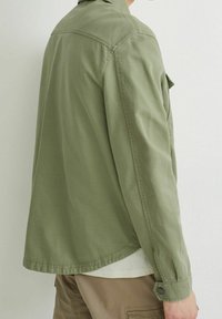 Keve olive green takki, jossa on napitettu hihansuu, rento leikkaus ja hieman kaareva helma, valmistettu sileästä, teksturoidusta kankaasta.