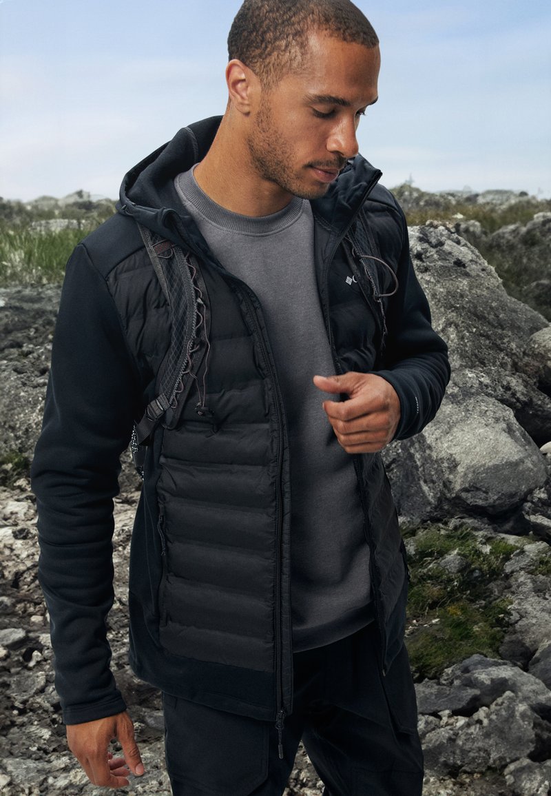 Black puffer jacket with hood, featuring quilted panels and zippered pockets. Worn over a gray crewneck shirt, set against rocky terrain.