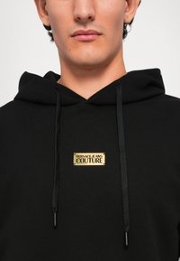 Homem a usar um hoodie preto com um logótipo dourado "Versace Jeans Couture" na zona do peito, visível do pescoço até à parte superior do tronco.
