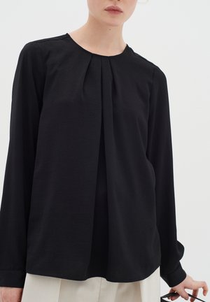 Vrouw die een zwarte blouse met lange mouwen en voorplooien draagt, houdt een zwartmontuur bril in haar rechterhand tegen een effen achtergrond.