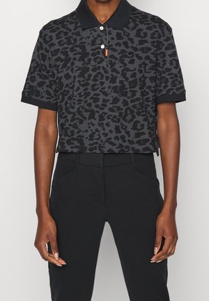Personne portant un polo à manches courtes noir et gris foncé à motif léopard et un pantalon noir, debout devant un fond uni.